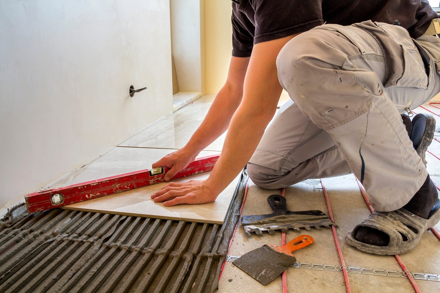 Young worker tiler installing ceramic tiles using lever on cement floor with heating red electrical cable wire system. Home improvement, renovation and construction, comfortable warm ،me concept.