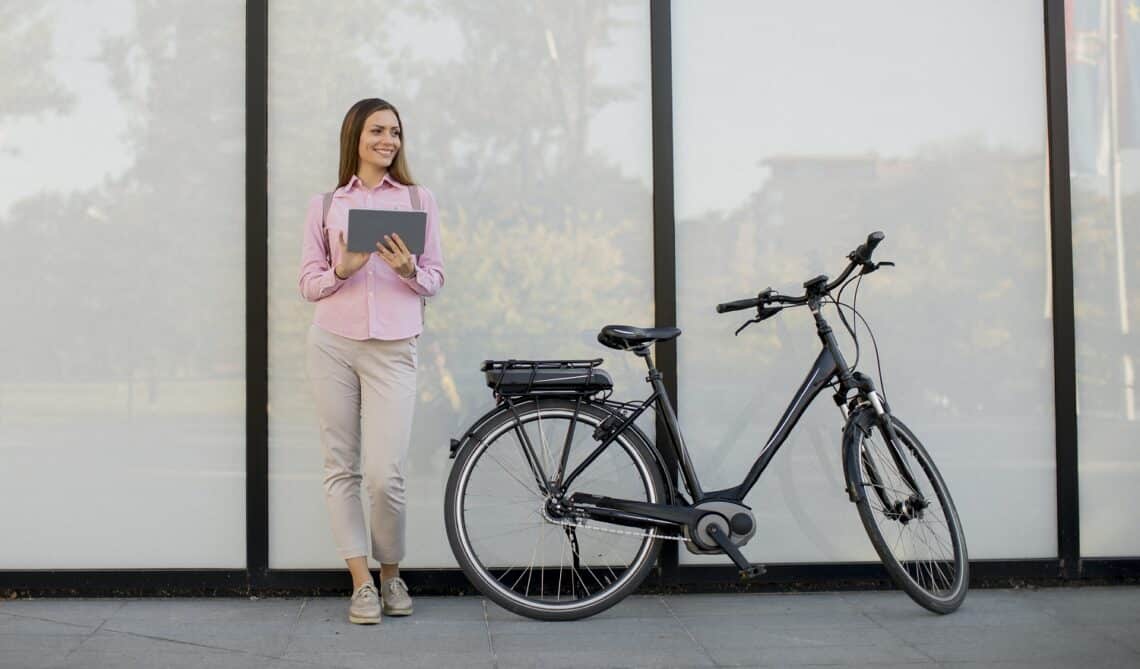 Pretty young woman standing by an electric bicycle and using digital tablet in urban environment