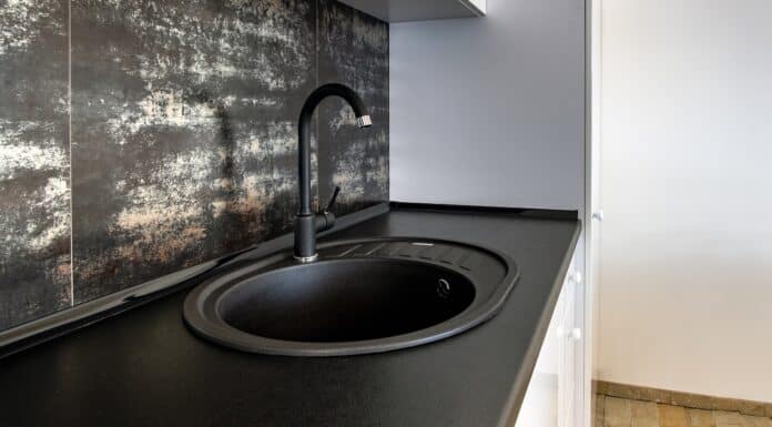 Interior of modern spacious kitchen with white contemporary furniture, black ceramic tiles on the wall and dark granite sink with water tap.