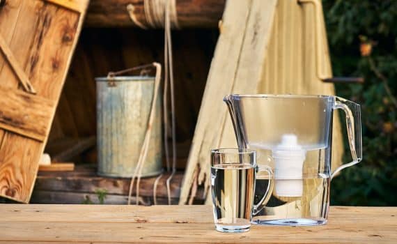 Water filter jug and a transparent glass cup of clean water in front of wooden draw well outdoors in summer evening at countryside