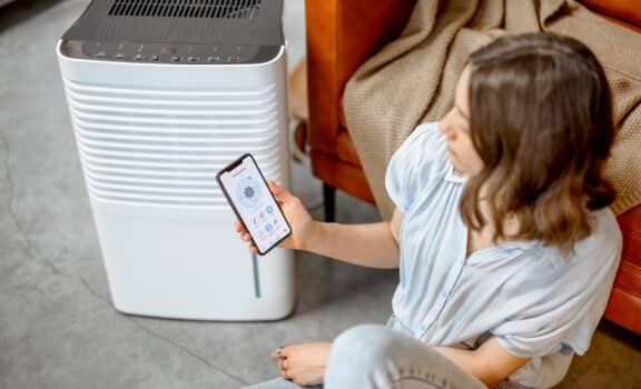 Pretty woman sitting near air purifier and moisturizer appliance near sofa monitoring air quality in phone. Health microclimate at home concept.