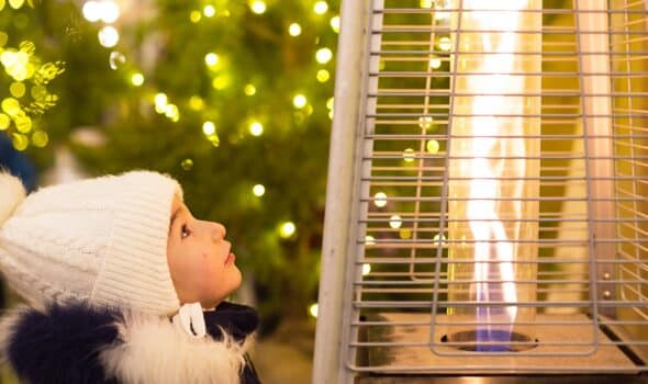 A little girl in a warm hat looks at the flame of a gas burner warming at a christmas market in the city. Lights of garlands in defocus on the christmas tree, new year, festive festivities
