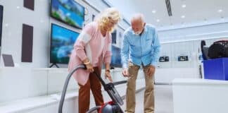 Cute smiling Caucasian senior couple trying out new vacuum cleaner for their home. Tech store interior.