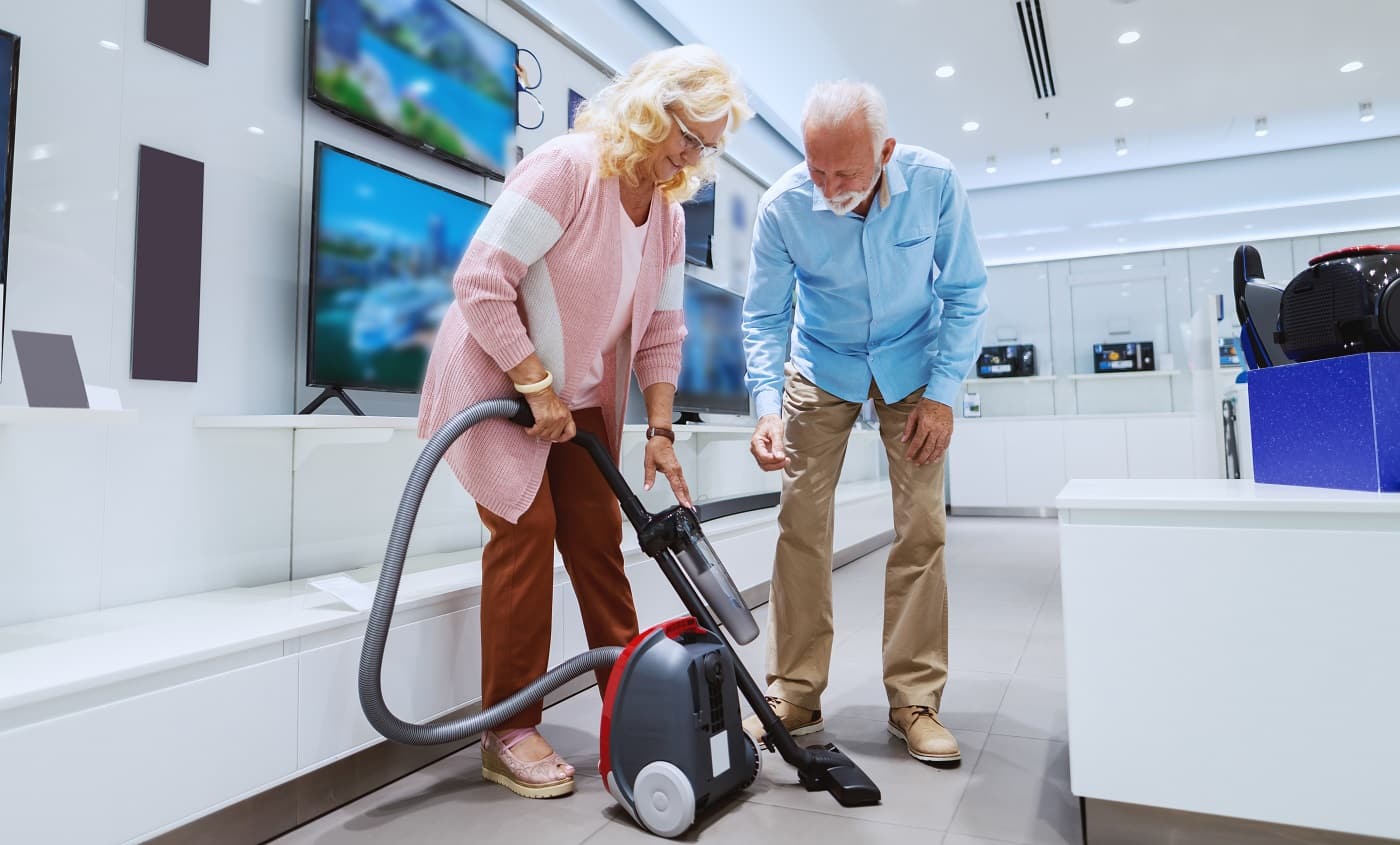 Cute smiling caucasian senior couple trying out new vacuum cleaner for their home. Tech store interior.