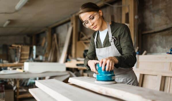 Assiduous young woodworker wearing safety glasses and apron while using electric sander in spacious workshop, waist-up portrait