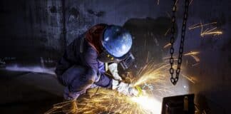 Male worker grinding on steel plate with flash of sparks close up wear protective gloves oil inside confined spaces.