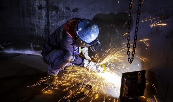 Male worker grinding on steel plate with flash of sparks close up wear protective gloves oil inside confined spaces.