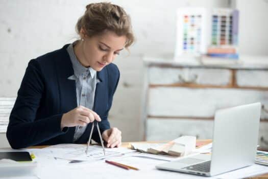 Portrait of beautiful young designer woman wearing suit using drawing compass on drafts. Attractive model working at office desk with blueprints for new project. Interior shot