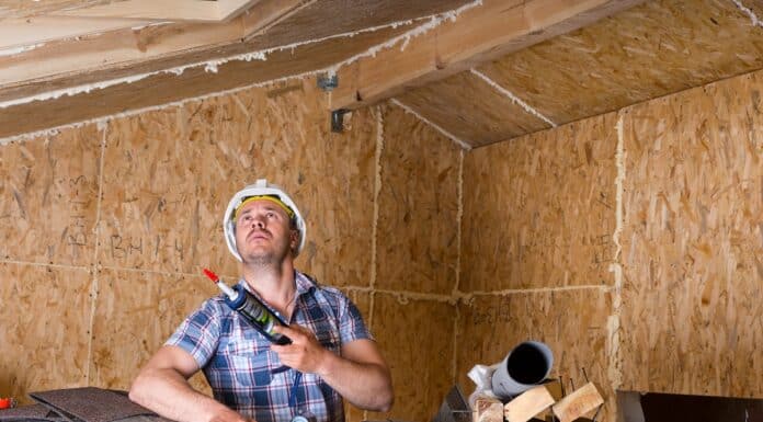 Male Construction Worker Builder Holding Caulking Gun and Looking Up at Ceiling Inside Unfinished Home with Exposed Particle Plywood Board