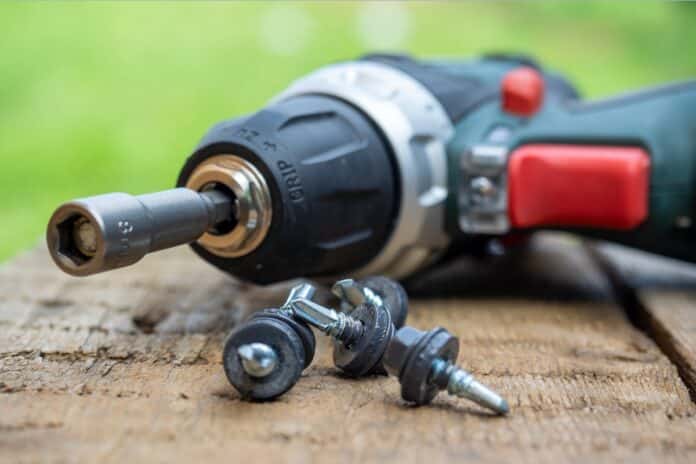 A close-up of an electric screwdriver with a nozzle on lies on a wooden background. Next to it, roofing screws on metal are laid out in a row. The concept of repair and construction