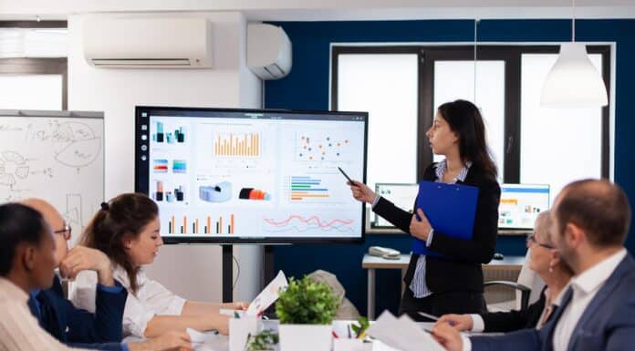 Young team leader in big corporation briefing coworkers pointing at graph meeting. Corporate staff discussing new business application with colleagues looking at screen