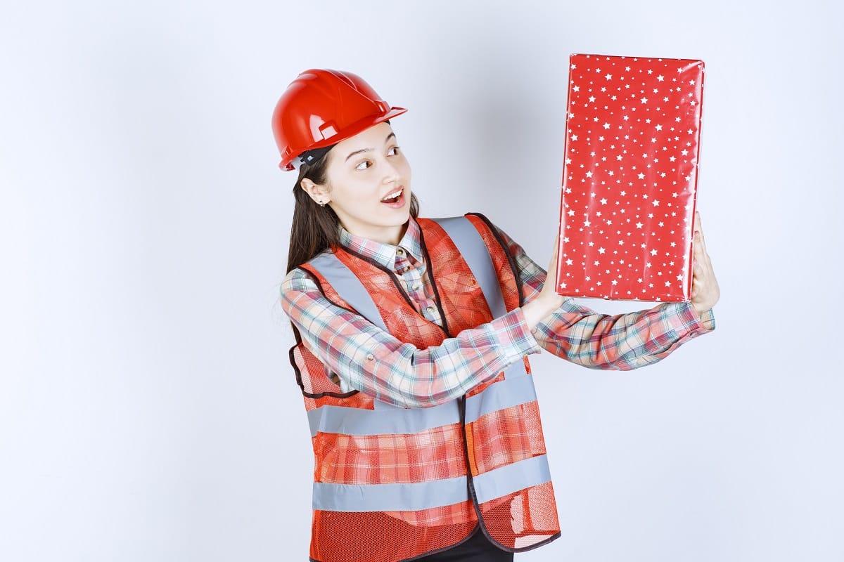 Young female engineer in red helmet looking at red gift box. High quality photo