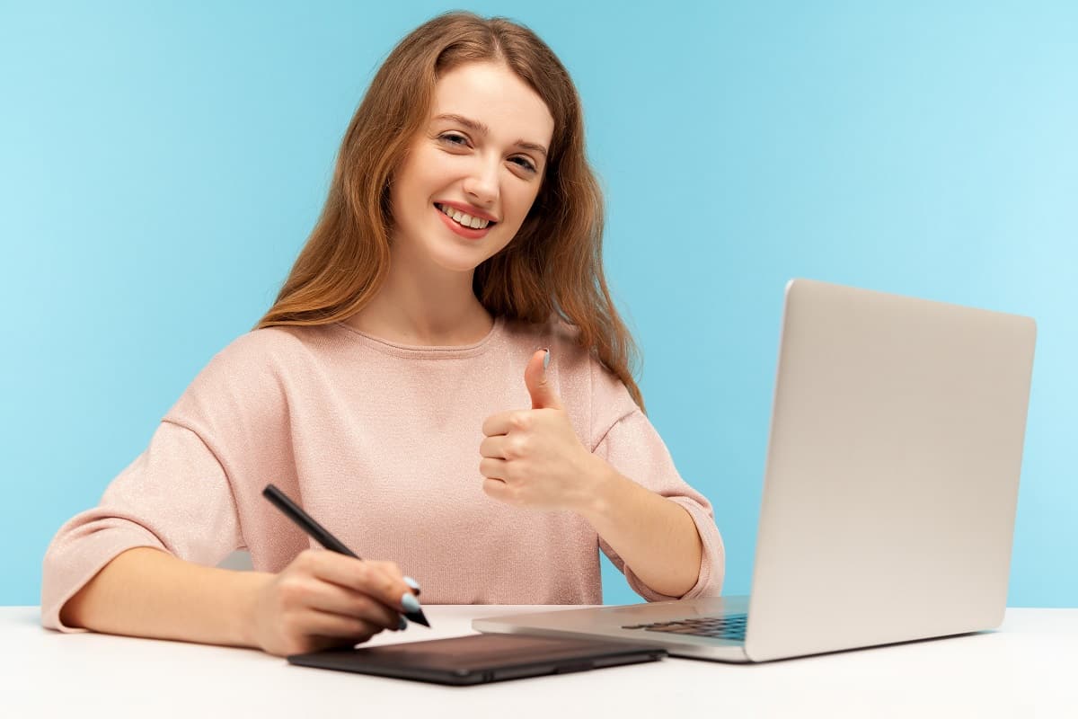 Smiling satisfied woman, creative designer sitting at workplace with graphic tablet and laptop, showing thumbs up while working on professional digital art equipment. Indoor studio shot, isolated