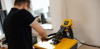 Man worker in glasses back using heat thermal textile application transfer machine. Printing application art design black T-shirt. Printing house office offset equipment. Manufacturing business label. How To Use Heat Press For T-Shirts.