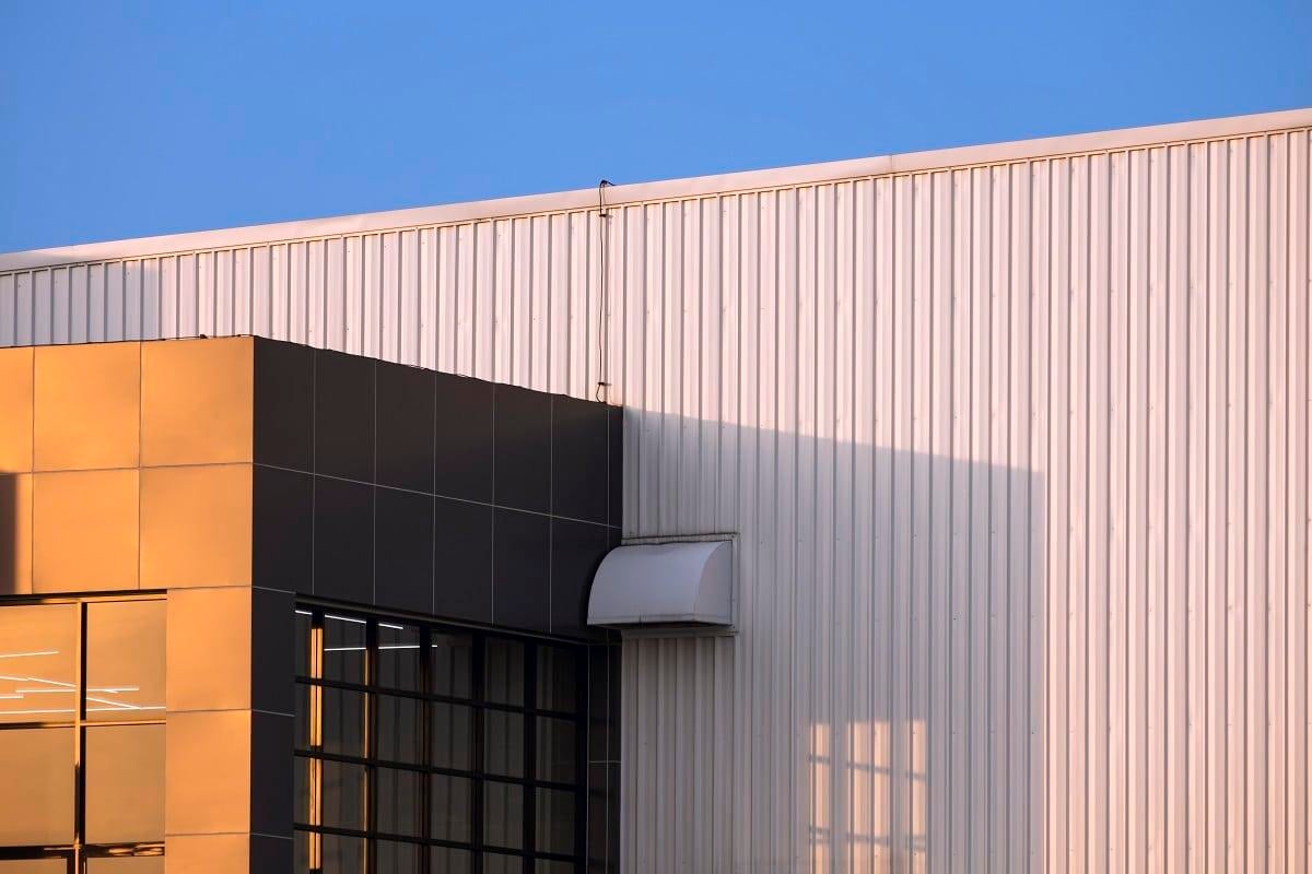 Sunlight on part of brown modern office on white corrugated metal wall of industrial building against blue sky background in evening time. Seamless steel log siding.