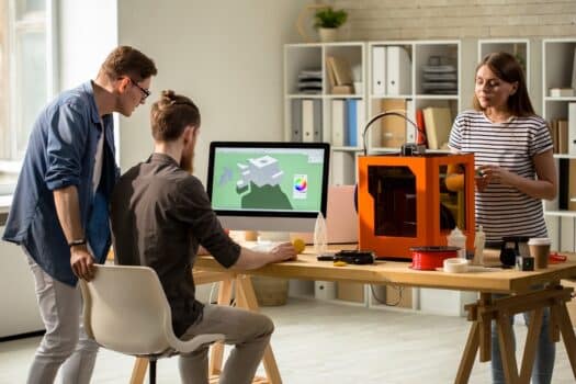 Back view portrait of young people working with 3d printer operating it via pc in modern design studio