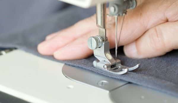 Hands of seamstress sewing with a professional machine, embroiders sews on grey fabric. Man seamstress sitting and sews on sewing machine in studio. Sewing workshop. Concept of factory, home, at work. How to make blackout curtains frequently asked questions.