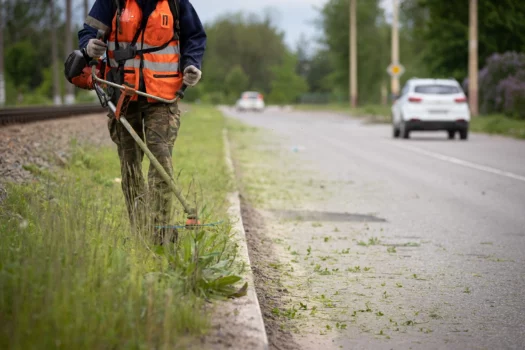 Front view of a worker in protective clothing with a petrol lawn mower on wheels walking on a lawn with grass along the roadway. A man mows grass with dandelions by the road in the city. Does stihl offer 4-cycle trimmers.