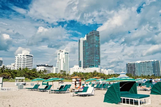 Miami, usa - january 10, 2016: sandy beach with people relaxing on deck chairs under green umbrellas, palms, cloudy blue sunny sky. Summer vacation. Lounge and leisure, miami beach or south beach. The perigon condos, miami beach - location.